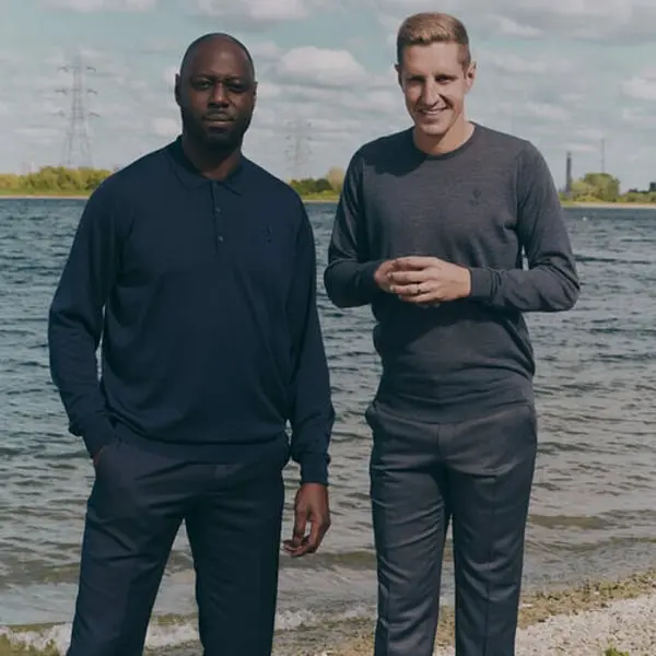 Deux hommes en tenues John Smedley posent au bord d’un lac en plein air, ciel partiellement nuageux.