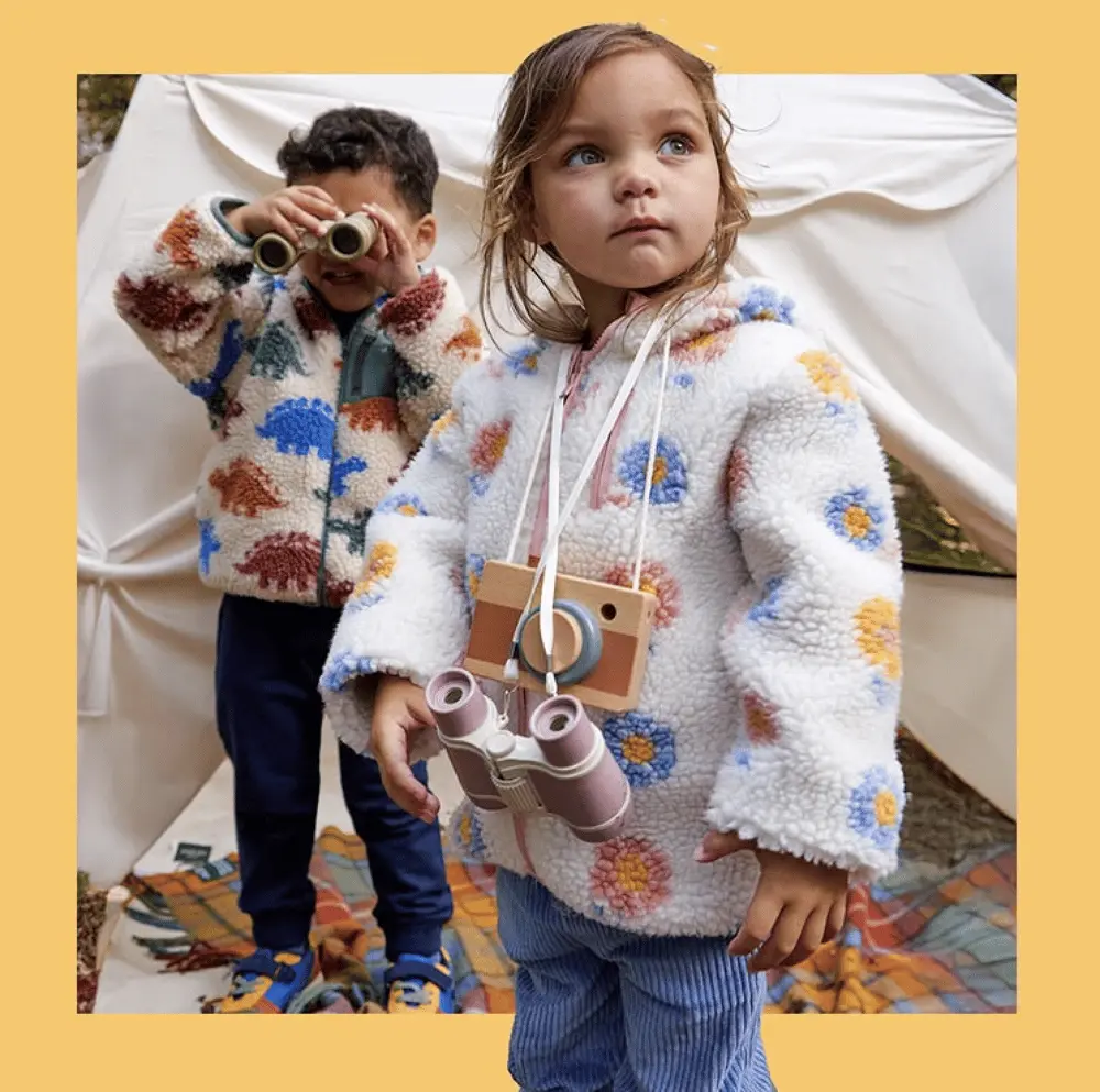 Deux enfants en vestes JoJo Maman Bébé avec jumelles et appareil photo jouets devant une tente crème