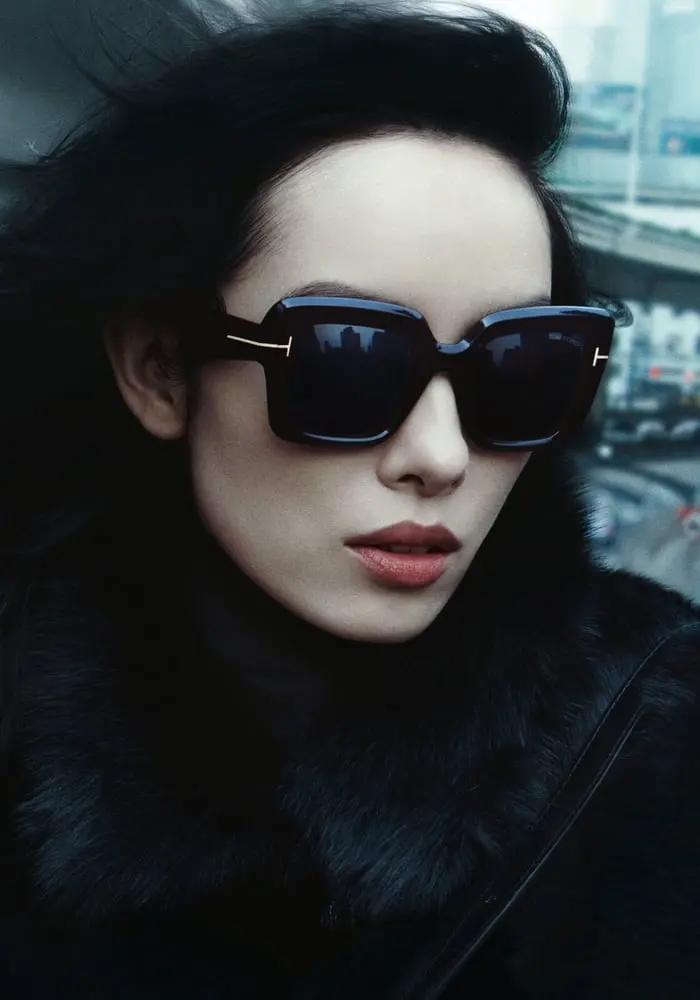 Personne portant des lunettes de soleil élégantes Marcolin, habillée d'un manteau en fourrure noire, décor urbain flou.