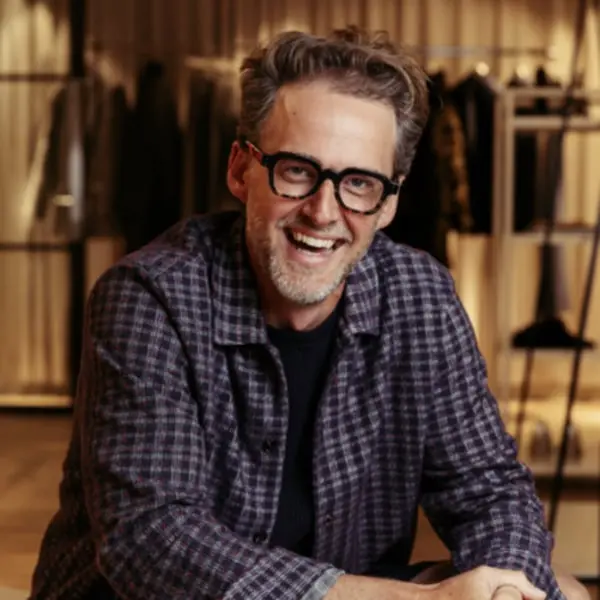 Un homme souriant en chemise à carreaux, fond flou de vêtements, évoquant Matalan, lumière chaleureuse