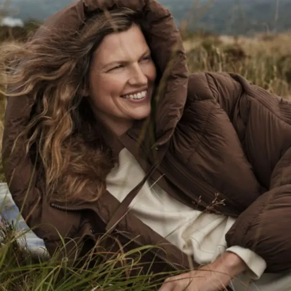 Personne souriante en manteau brun dans un champ venteux, illustrant la collection automnale Matalan