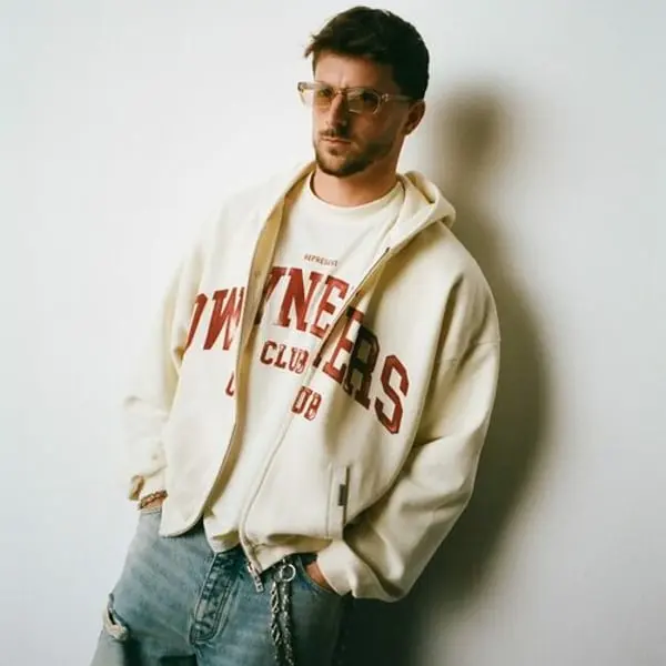 Individu en sweat à capuche "Owners Club" blanc de Represent, lunettes de soleil, jeans décontractés, pose décontractée.