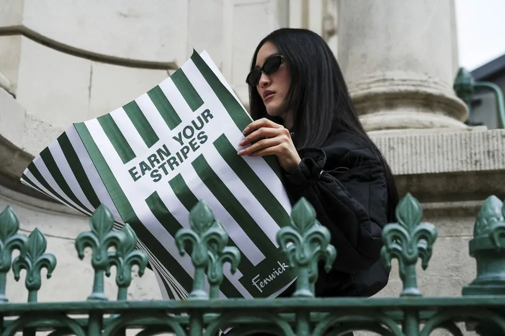Femme lisant une affiche "Earn Your Stripes" de Fenwick devant une structure architecturale ornée de motifs floraux