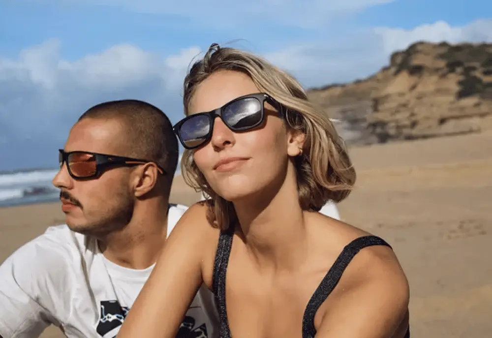 Deux personnes portent des lunettes de soleil Safilo sur une plage ensoleillée avec des falaises en arrière-plan
