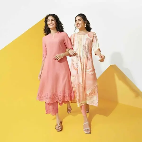 Deux femmes souriantes en robes colorées de Shoppers Stop marchent ensemble devant un fond géométrique jaune.