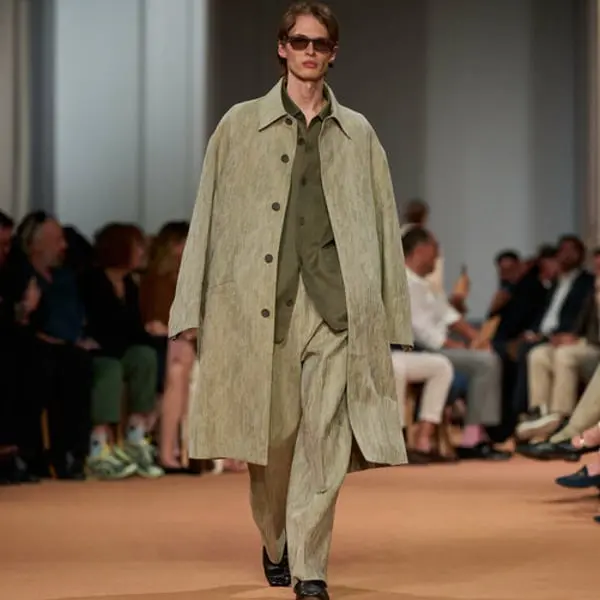 Un mannequin défile en tenue Zegna beige sur un podium, avec des spectateurs en arrière-plan.