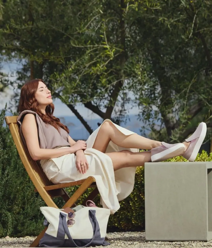 Une femme détendue porte des chaussures blanches Allbirds, assise sur une chaise en bois dans un jardin verdoyant.