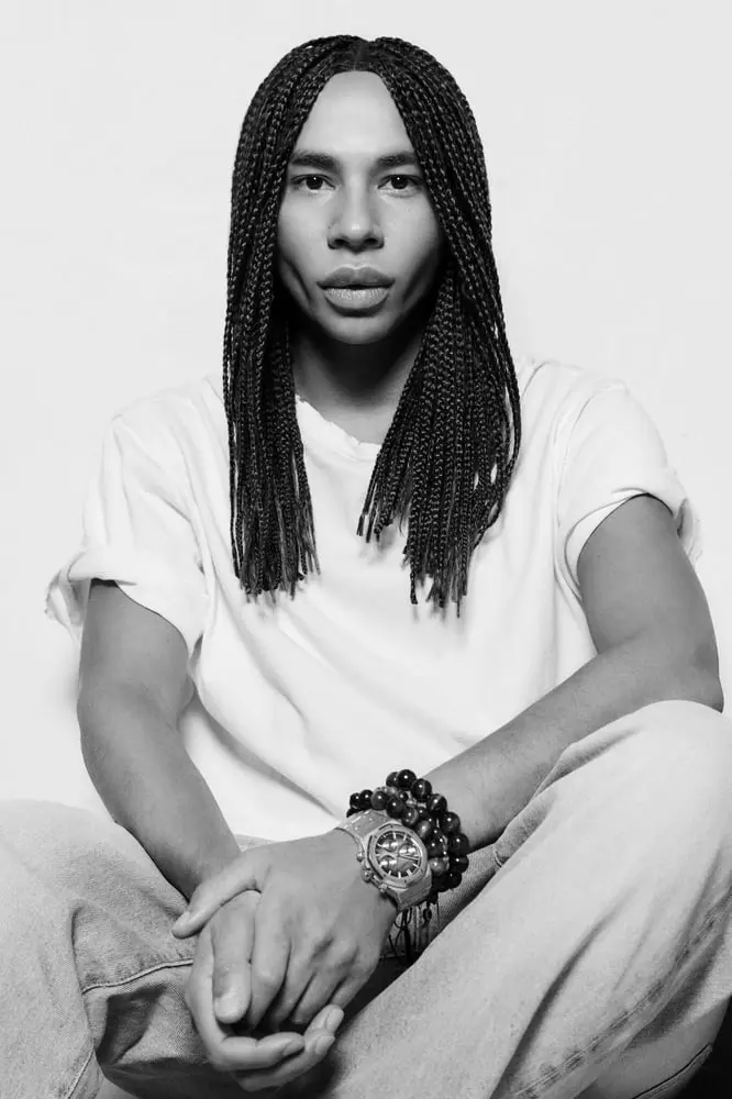 Olivier Rousteing assis, vêtu d'un t-shirt blanc et de bracelets, devant un fond neutre, représentant Balmain