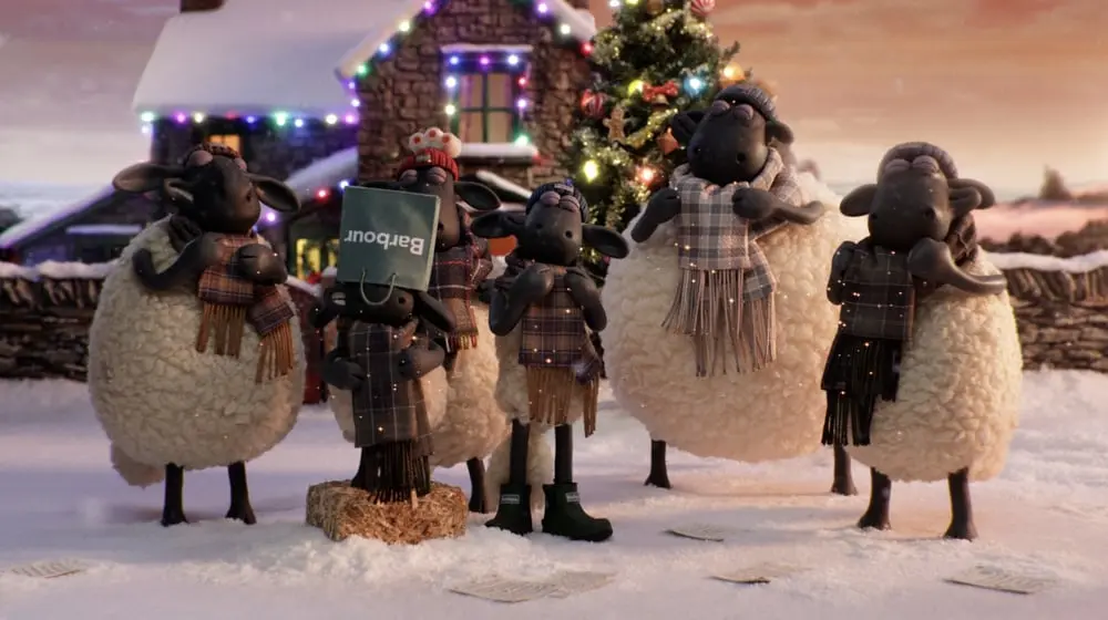 Des moutons vêtus de foulards devant un sapin de Noël illuminé et une maison, l'un porte un sac marqué Barbour