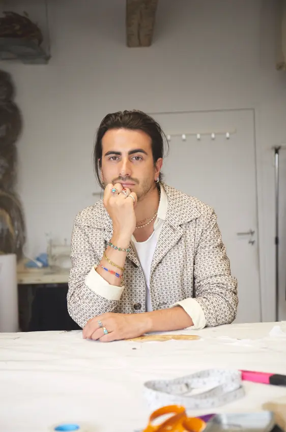 Homme assis pensif dans un atelier de couture, portant une veste chic et entouré d'outils de création Benmoyal