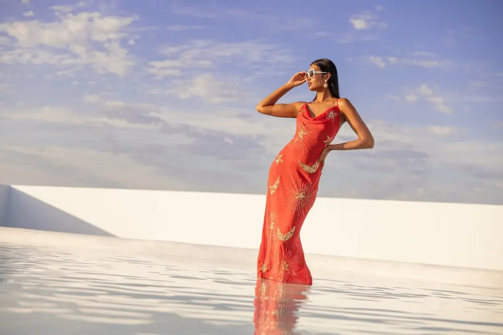 Femme en robe rouge à motifs, debout au bord d'une piscine sous un ciel bleu, évoquant le style de Debenhams