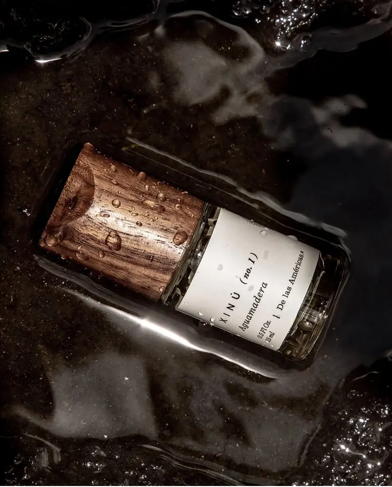 Flacon de parfum Xinú avec bouchon en bois, posé sur une surface humide avec des reflets d'eau.