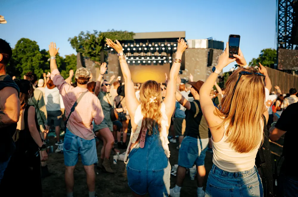 Des fans de concert lèvent les bras et prennent des photos devant une scène en plein air, vêtus de tenues d'été.