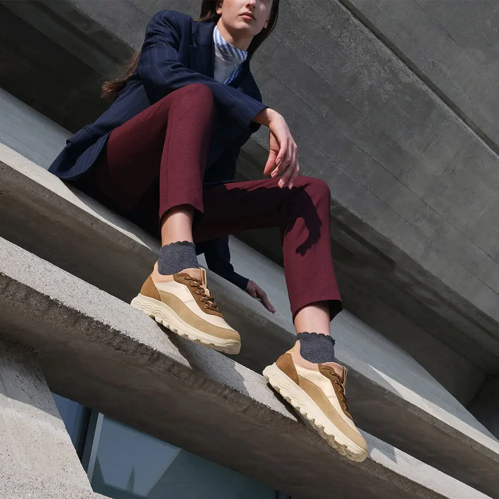 Personne portant une veste sombre assise sur un béton avec des baskets Geox marron-beige