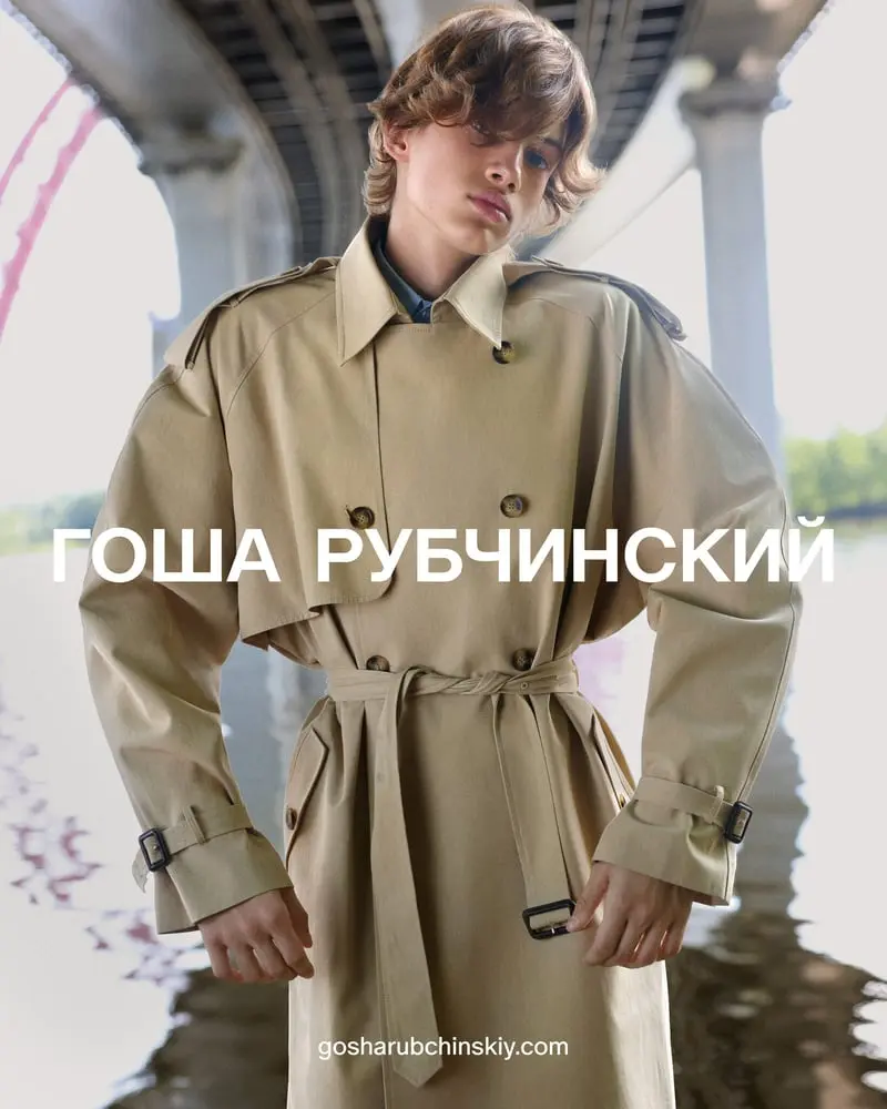 Un mannequin en trench-coat beige pose sous un pont avec le logo Gosha Rubchinskiy en blanc