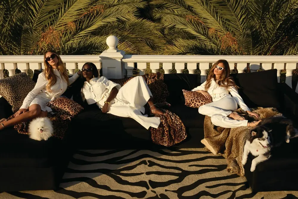 Trois femmes en tenue blanche et lunettes de soleil posent sur un canapé noir, entourées de motifs léopard et de chiens.