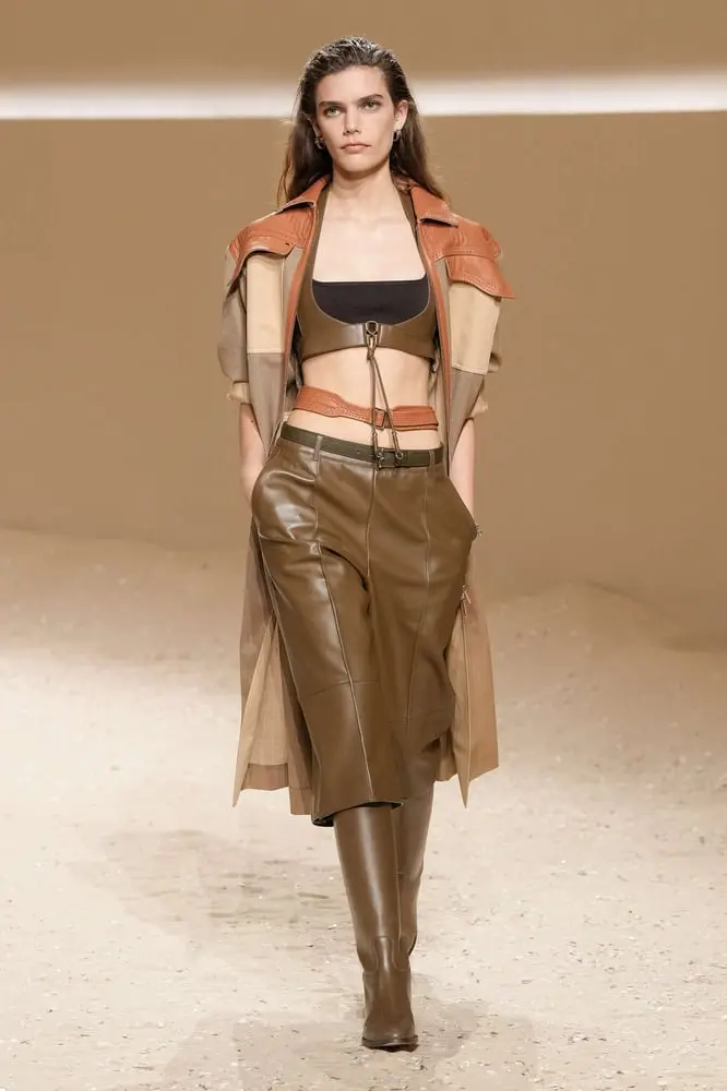 Mannequin défilant en tenue en cuir Hermès, jupe et bottes marron, sur un podium beige