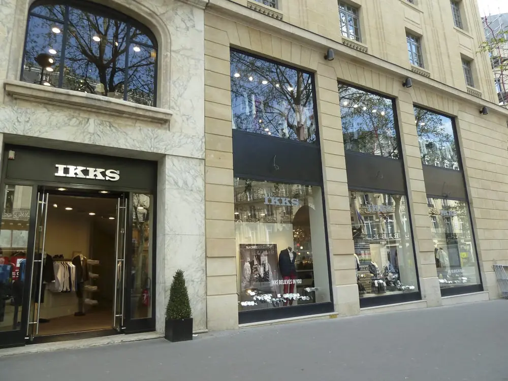 Boutique IKKS dans un bâtiment en pierre avec de grandes vitrines affichant des vêtements et un logo visible