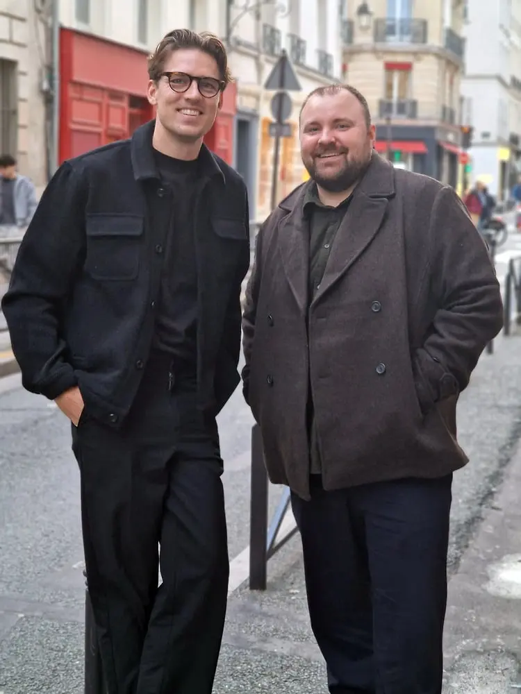 Deux hommes souriants en manteaux foncés posent dans une rue parisienne proche de la boutique Les Deux