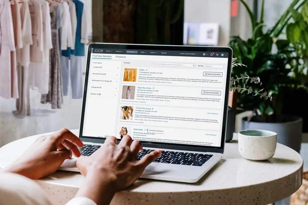 Utilisateur recherchant des détaillants de mode sur le site Joor, ordinateur portable sur table avec vêtements en arrière-plan