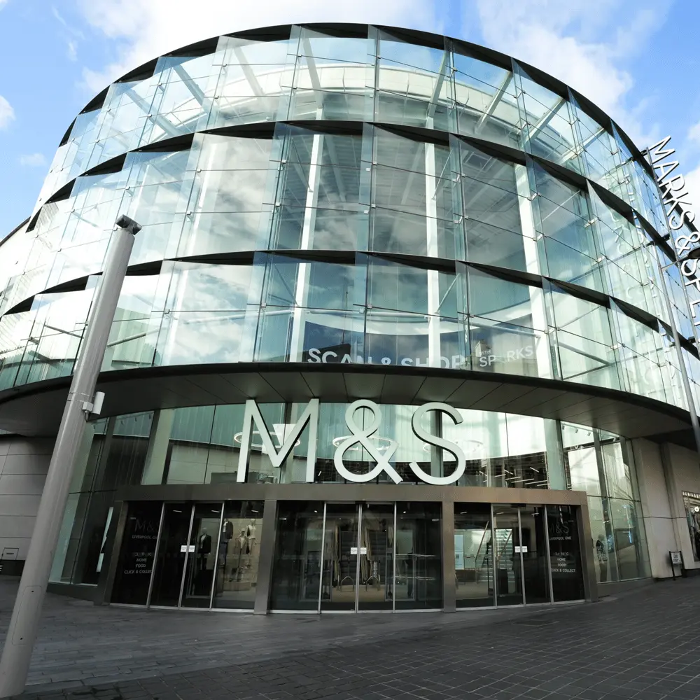Entrée du magasin M&S avec une façade en verre moderne sous un ciel nuageux.