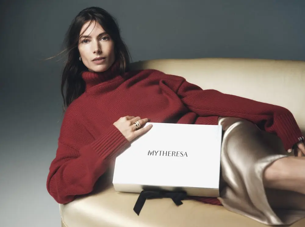 Femme en pull rouge tenant une boîte Mytheresa sur un canapé beige, reflétant une image de luxe et de confort
