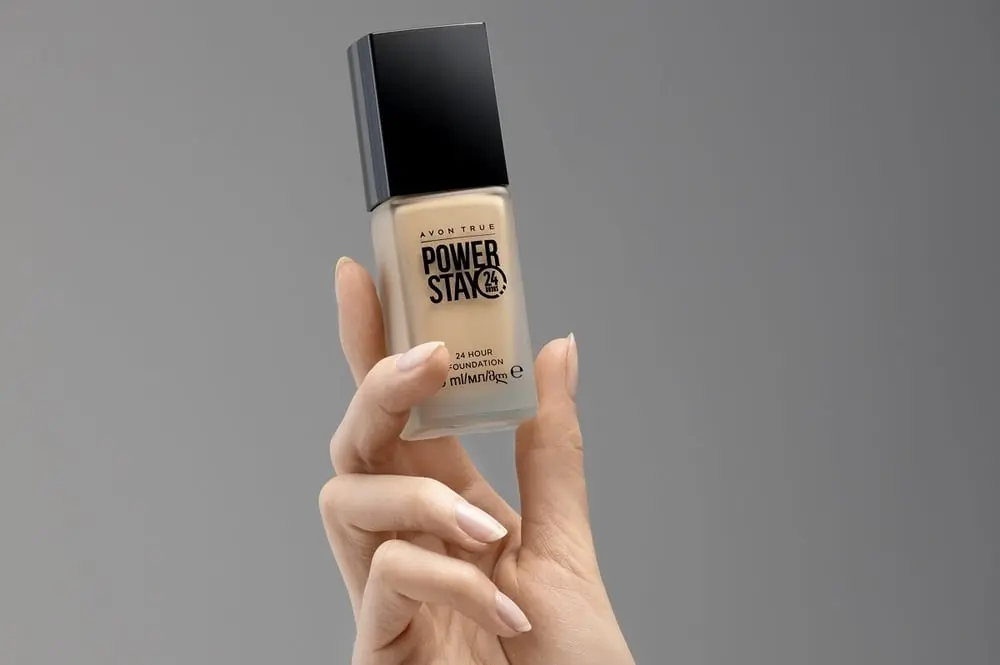 Main tenant un flacon de fond de teint Avon True Power Stay sur fond gris
