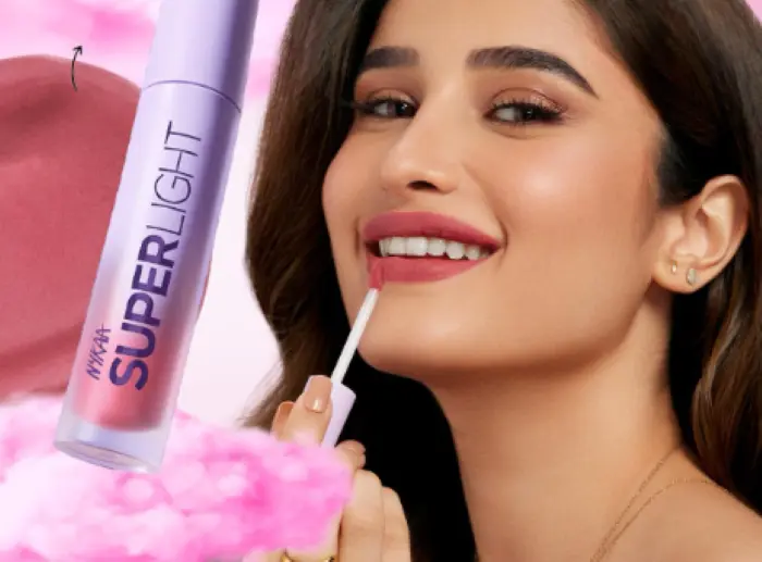 Une femme souriante applique un gloss Nykaa SuperLight avec des fleurs roses en arrière-plan.