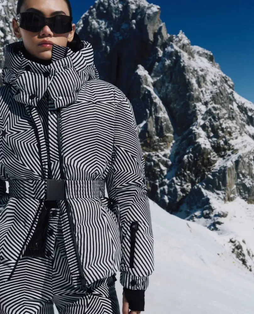 Personne en vêtement de ski Perfect Moment rayé en noir et blanc, devant un paysage montagneux enneigé.
