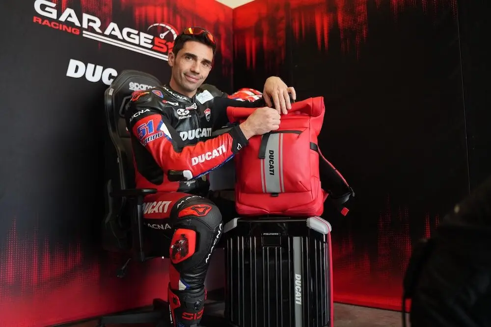 Pilote en combinaison Ducati posant avec un sac à dos rouge Ducati dans un garage d'écurie de course.