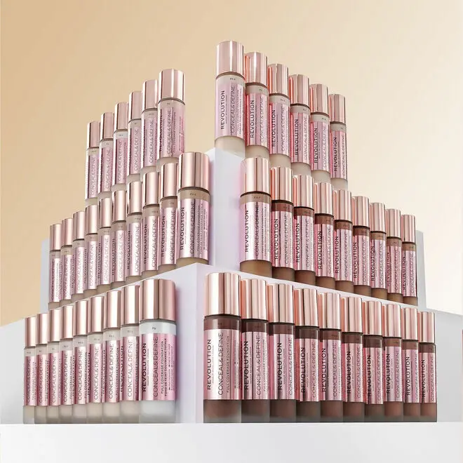 Flacons de maquillage Revolution Beauty alignés en pyramide sur fond beige, étiquettes roses et bouchons dorés.