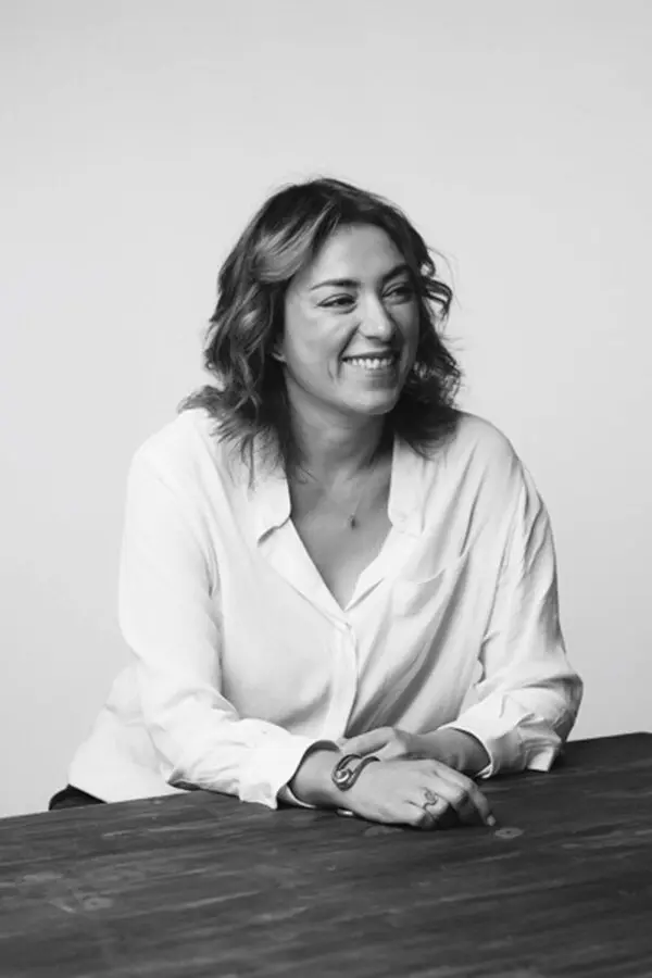Une femme souriante en chemise blanche est assise à une table en bois, associée à la marque Ba&sh