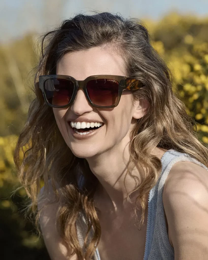 Une femme souriante portant des lunettes de soleil, entourée de végétation luxuriante, pour Safilo