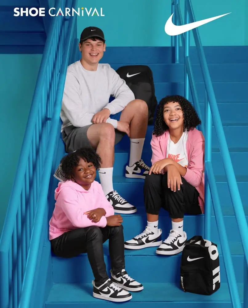 Trois enfants assis sur des escaliers bleus portant des articles Nike et souriant, logo Shoe Carnival en haut à gauche