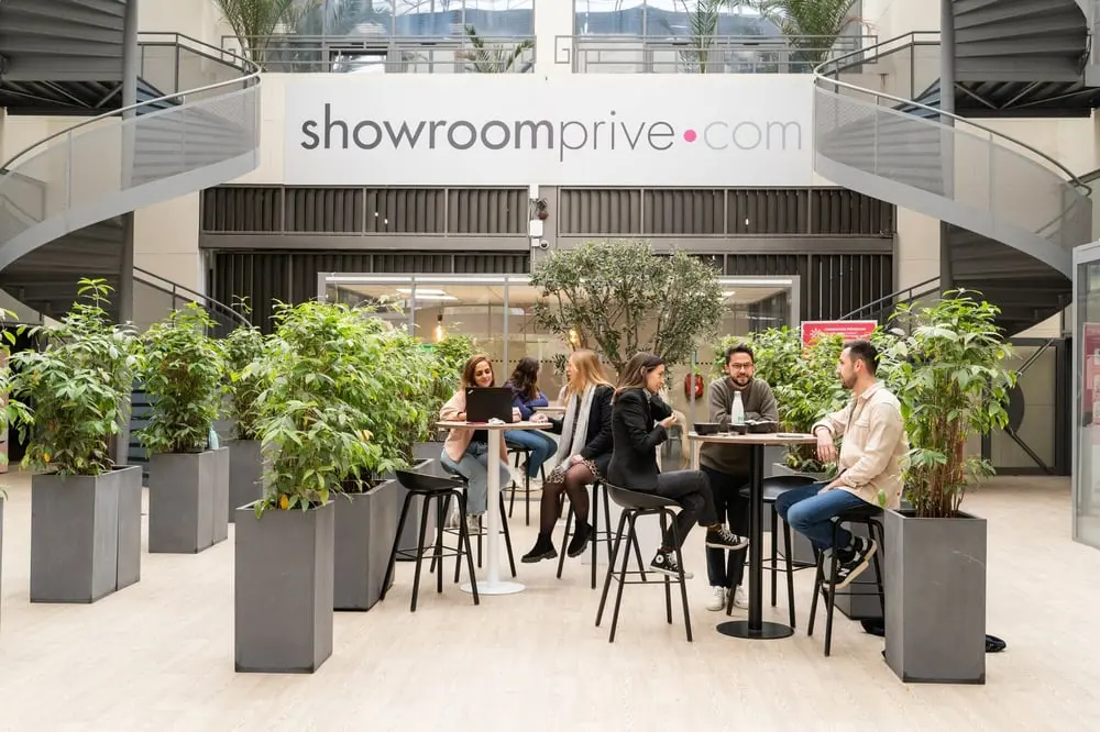 Des personnes discutent autour de tables dans un espace moderne où le logo Showroomprivé est visible en arrière-plan