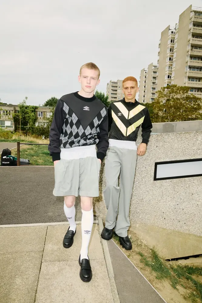 Deux jeunes hommes en vêtements Umbro posent devant des immeubles, combinant style rétro et moderne.