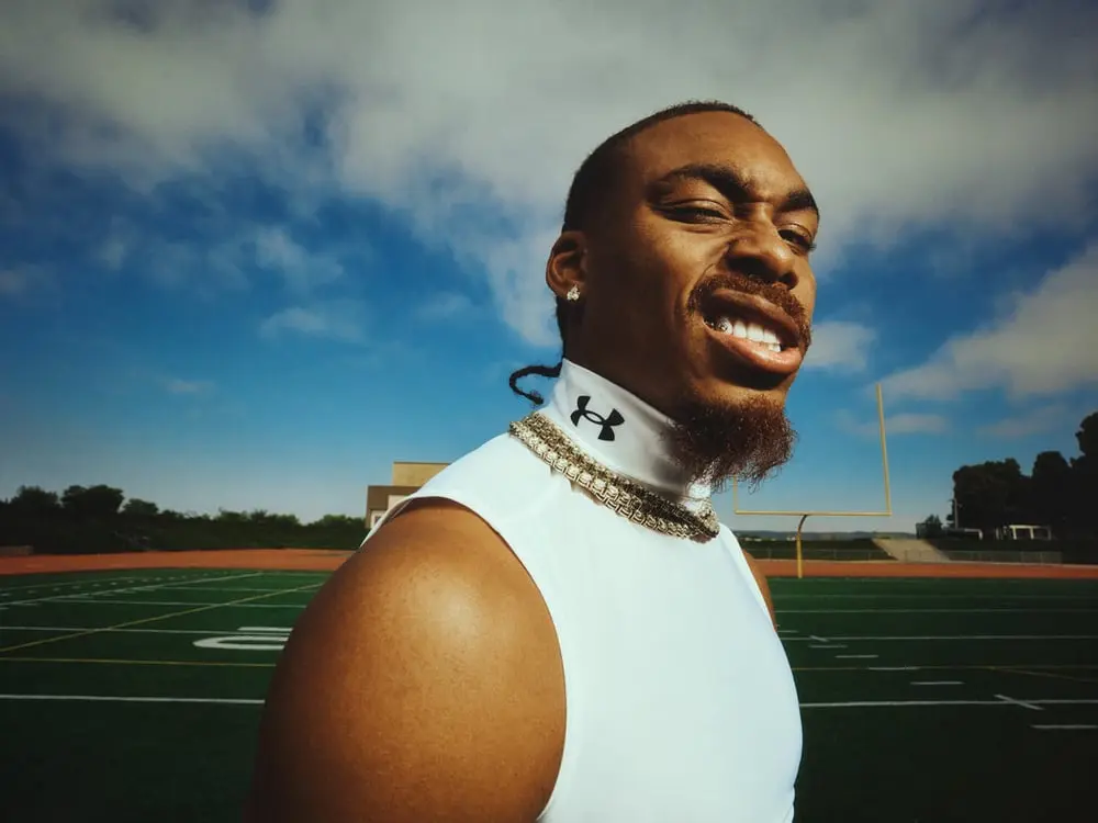 Un homme en tenue de sport Under Armour sourit sur un terrain de football sous un ciel partiellement nuageux