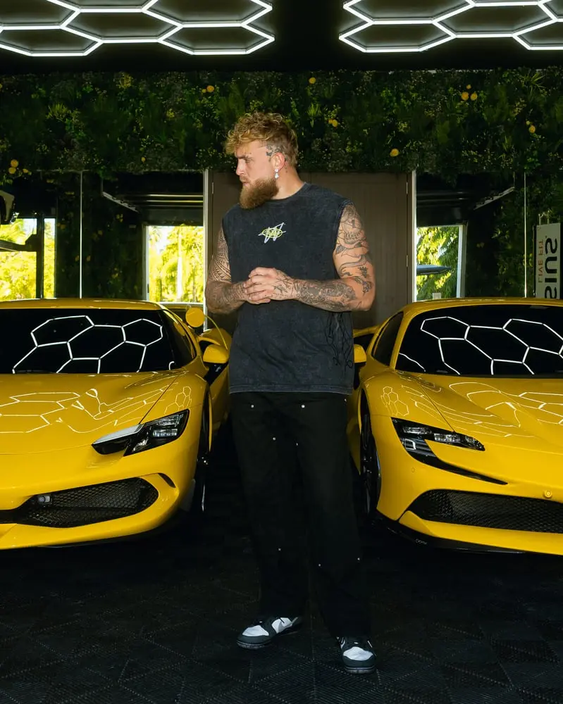 Homme tatoué debout entre deux voitures jaunes avec un logo W, éclairage moderne, plantes en arrière-plan