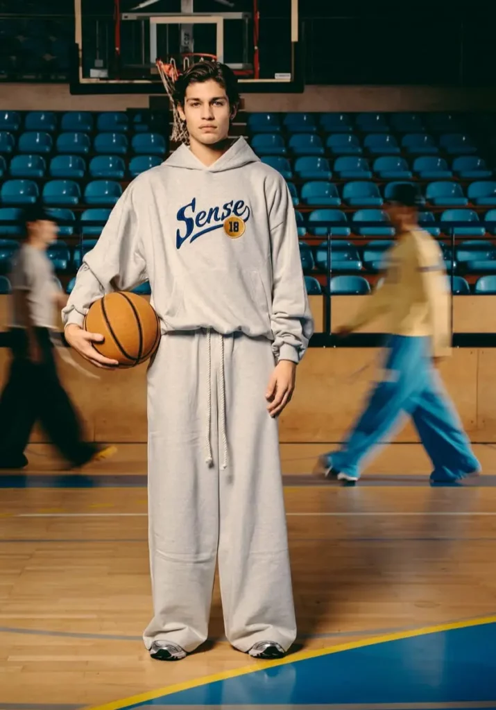 Un mannequin en survêtement gris Sense 18 tient un ballon de basket sur un terrain avec des sièges bleus en arrière-plan