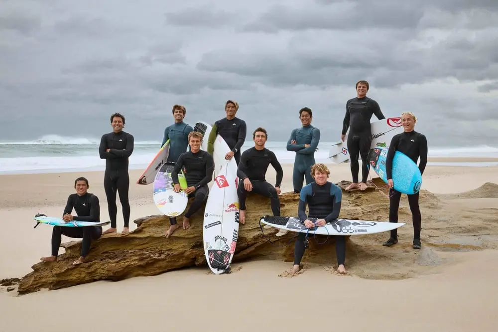 Groupe de surfeurs en combinaisons Quiksilver et RVCA posant sur une plage avec des planches de surf