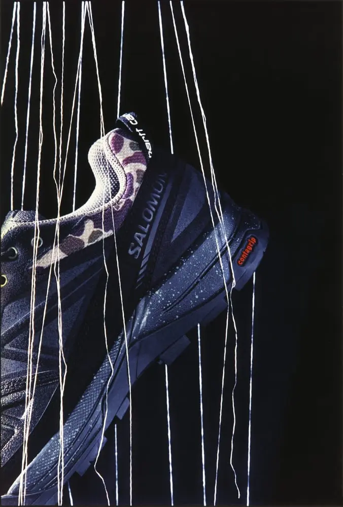 Chaussure Salomon au motif camouflage suspendue par des fils, illustrant une collaboration mode et technicité.