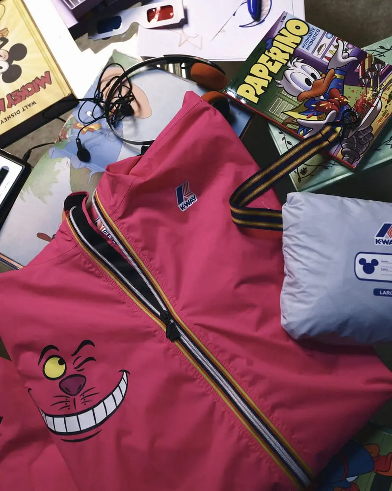 Veste rose K-Way avec le sourire du Chat de Cheshire, casque audio et bande dessinée Disney sur une table