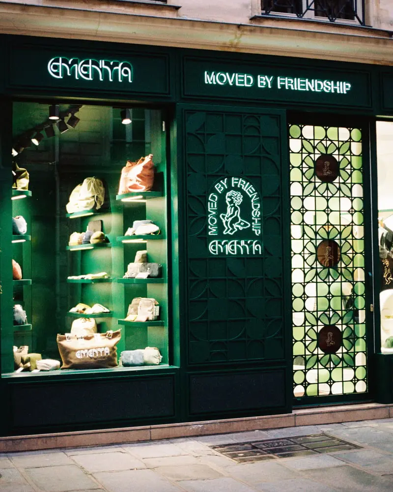 Façade verte de la boutique Ementa à Paris avec sacs et slogan "Moved by Friendship" en néon.