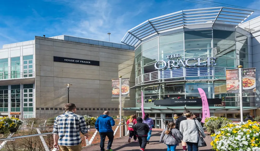 Passants devant l'entrée du centre commercial The Oracle à Reading sous un ciel bleu, avec le logo visible