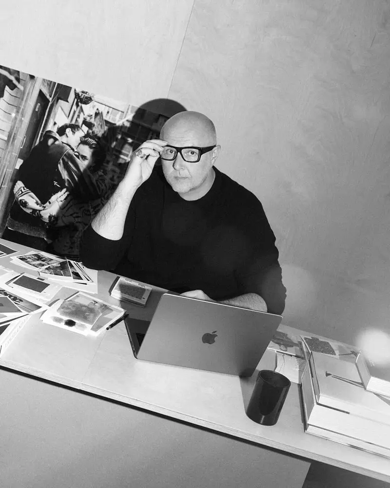 Homme chauve à lunettes assis à un bureau, posé devant des photos et un ordinateur portable Apple lors de la Fashion Week de Londres