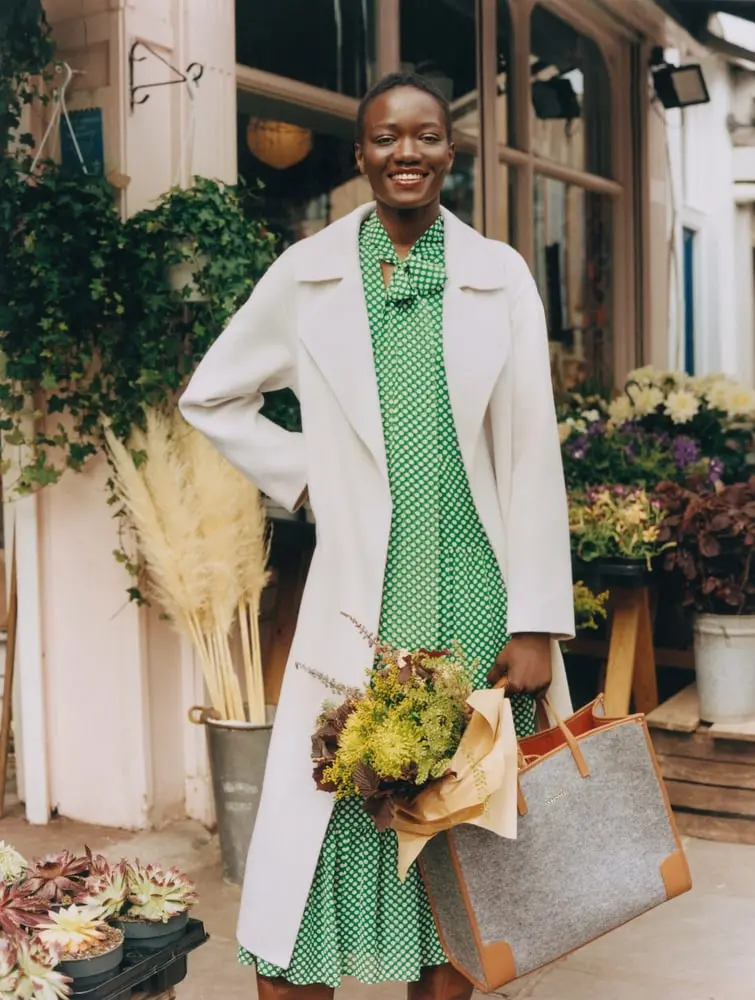 Une femme souriante en robe verte et manteau blanc, tenant un bouquet, devant un magasin de LK Bennett