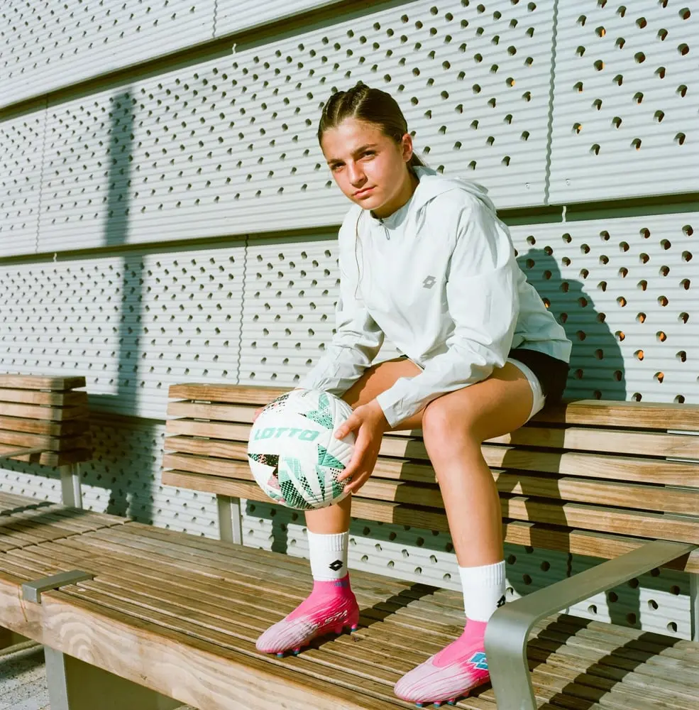 Jeune femme assise sur un banc avec un ballon Lotto, vêtue de chaussures roses, devant un mur perforé en métal