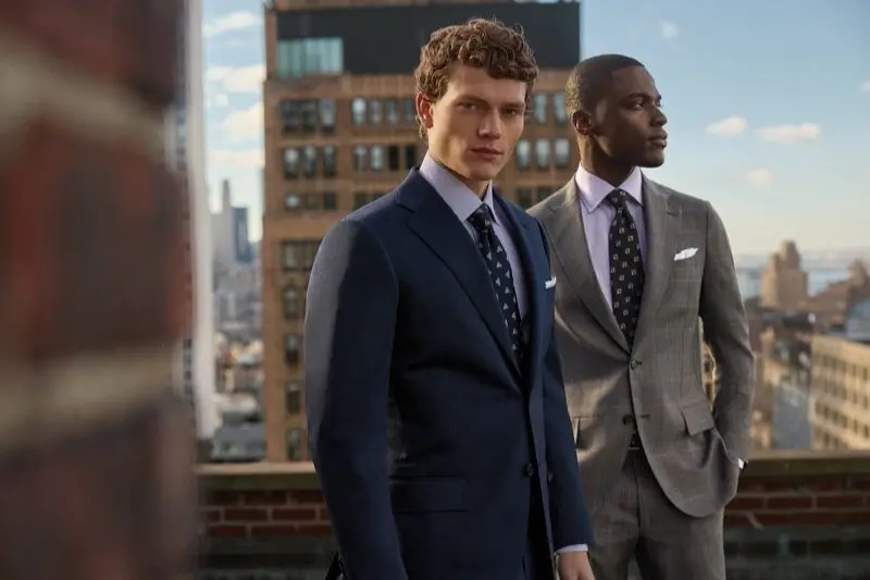 Deux hommes en costumes élégants Paul Stuart posent sur un rooftop avec vue sur des bâtiments urbains.