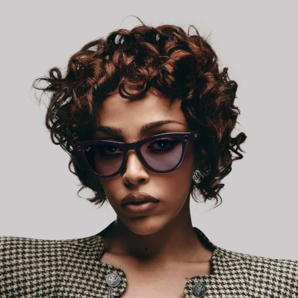 Femme portant des lunettes de soleil Ray-Ban Meta avec monture sombre, coiffure bouclée et veste à motifs.