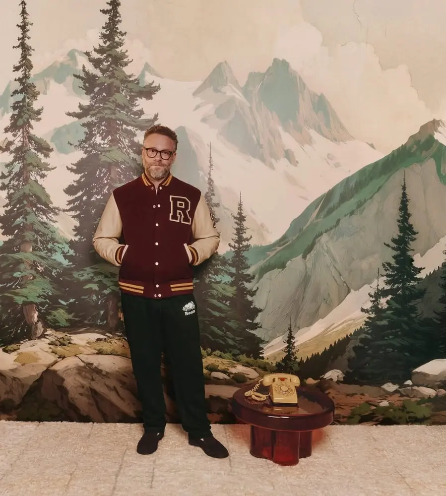 Un homme portant une veste à l’emblème de Roots pose devant un paysage de montagne peint.